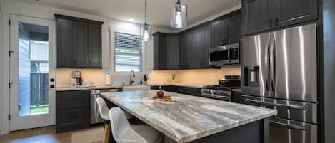 A modern kitchen with dark cabinets, stainless steel appliances, a marble island, and a pendant light.