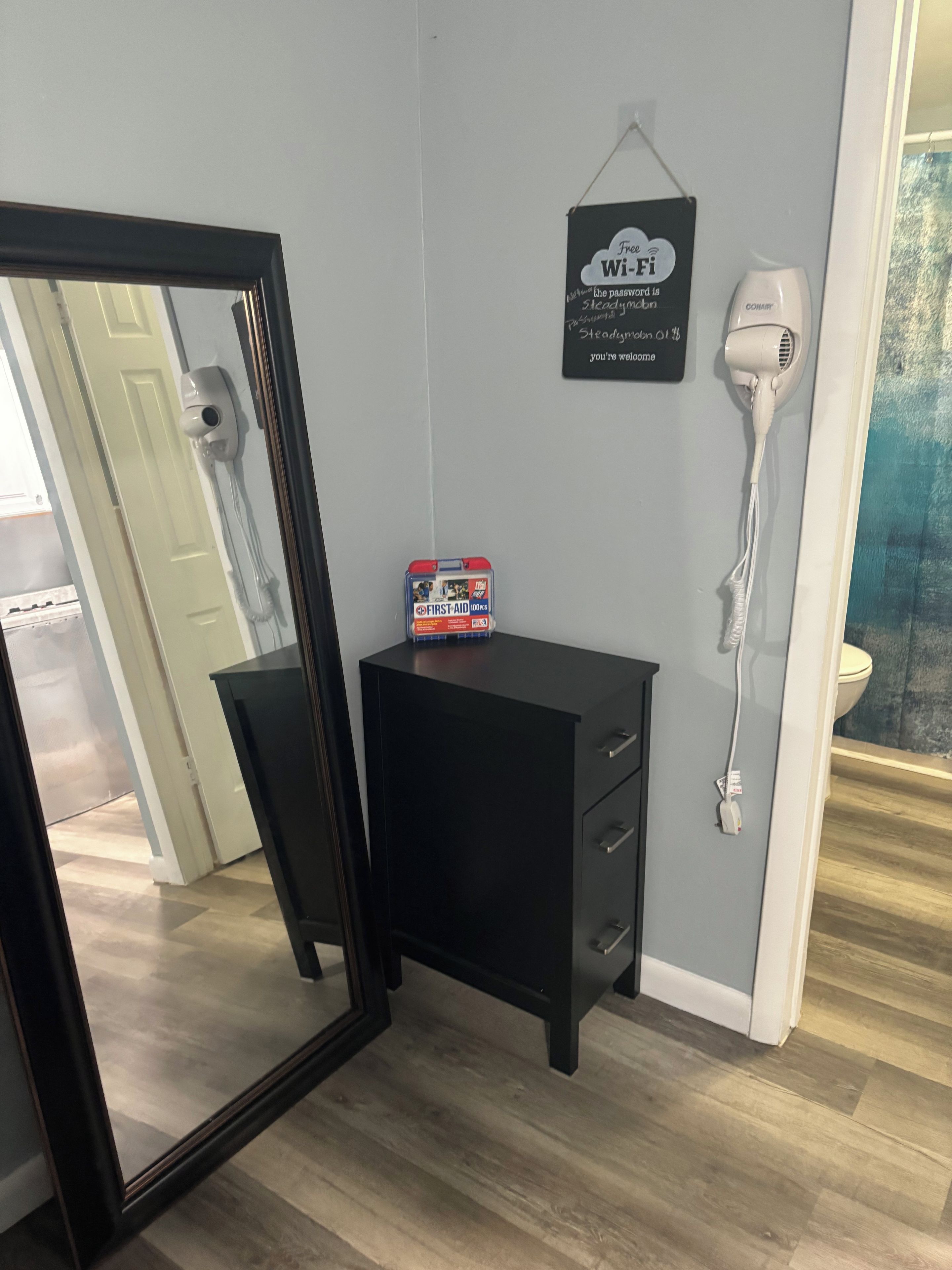 Une pièce comprenant une commode noire, un miroir, une trousse de premiers secours, un panneau indiquant la présence d’une connexion Wi-Fi et un sèche-cheveux.