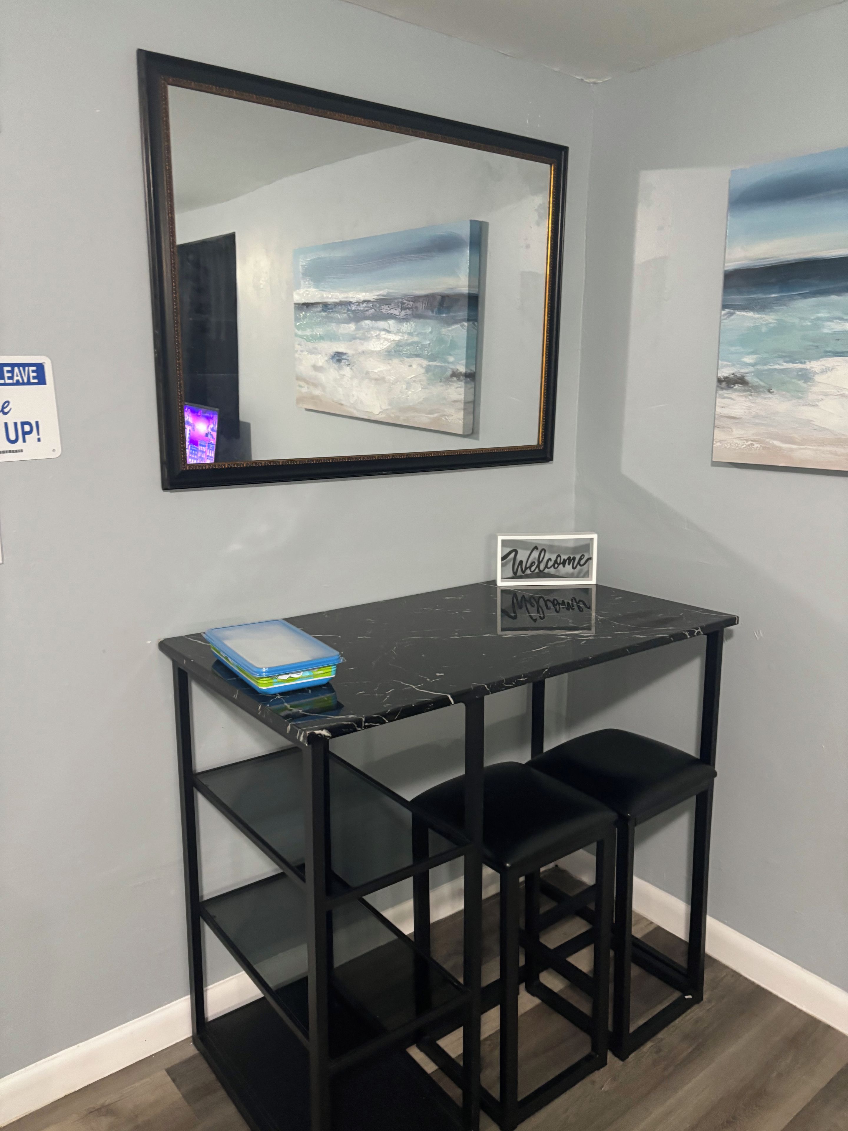 Un bureau en métal noir avec une pancarte « Bienvenue », deux tabourets de bar noirs, un tableau encadré représentant une plage et une pancarte « NE PAS ENLEVER !» accrochée au mur.
