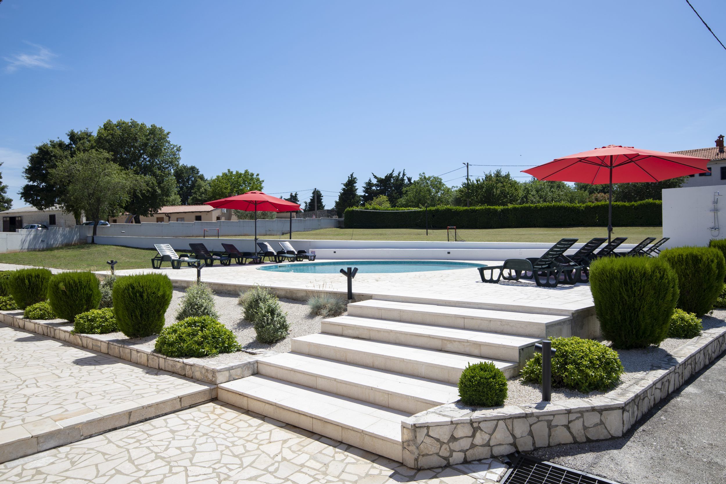 A pool area with lounge chairs and umbrellas, surrounded by well-maintained hedges and a stone pathway.