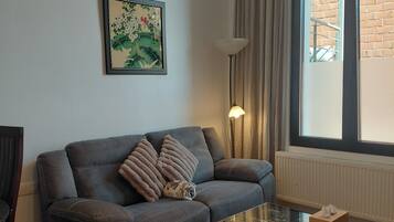 A living room with a grey sofa, a glass coffee table, and a large window with curtains.
