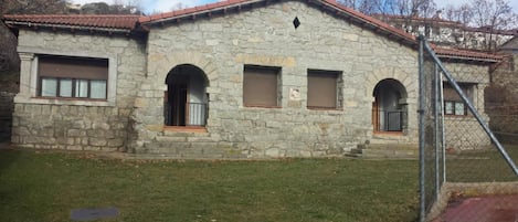 Un edificio de piedra con techo de tejas rojas, un pequeño jardín delantero y una cerca de metal.