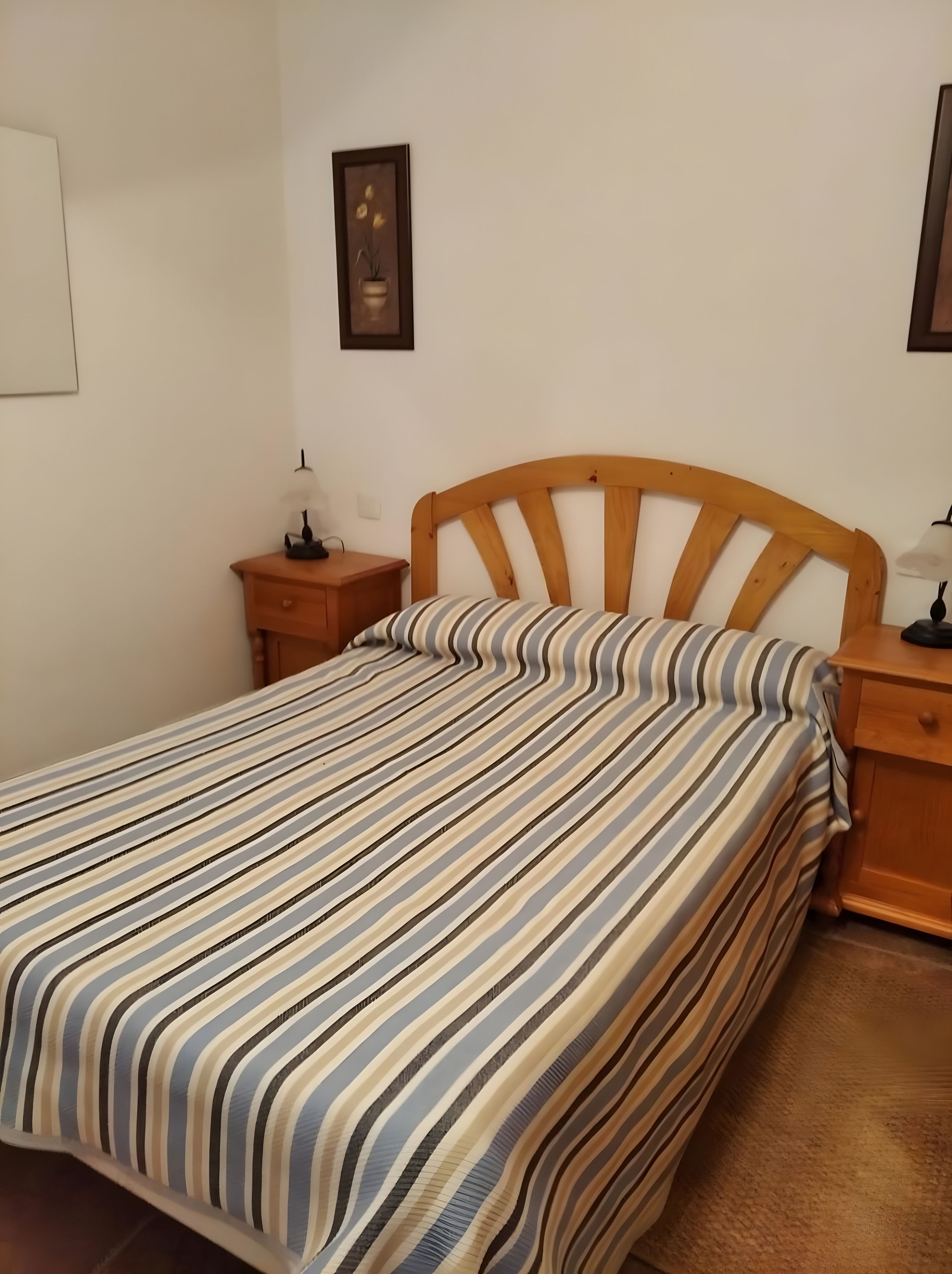 Una cama individual con una colcha a rayas, un cabecero de madera, una mesita de noche con una lámpara y un cuadro en la pared.