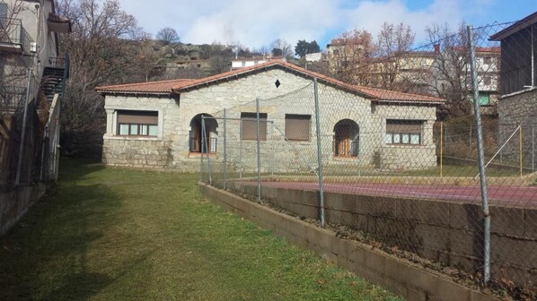 A stone building with a red-tiled roof, a fenced yard, and a hilly backdrop.