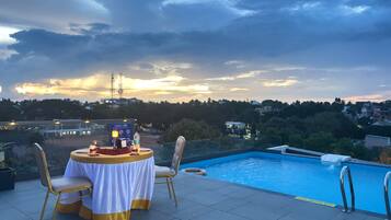 Un área de piscina en la azotea con una mesa y sillas, un letrero que dice 'HAVE FUN' y vistas de la ciudad al fondo.
