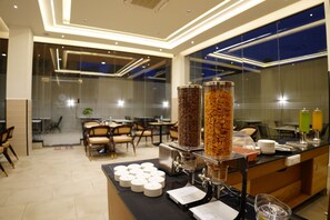 A modern hotel breakfast area with a self-serve station, including coffee and cereal dispensers, and a seating area with tables and chairs. - Crescent Hotel (Mysuru)