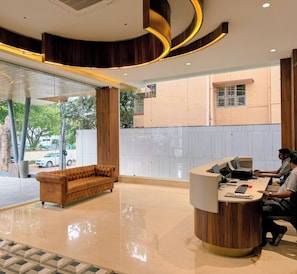 A reception area with a wooden reception desk, a brown leather sofa, and a tiled floor. - Crescent Hotel (Mysuru)