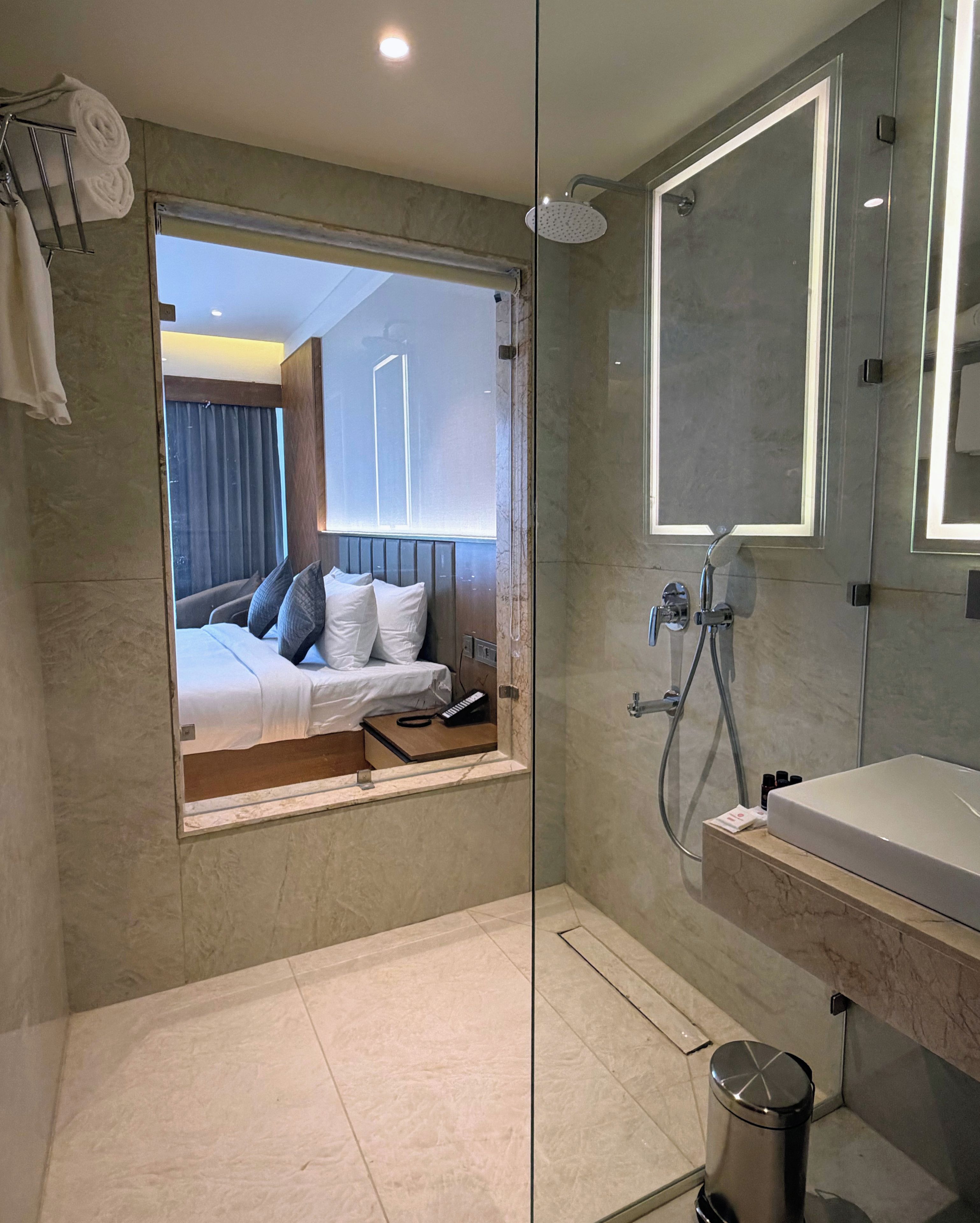 Un baño moderno con cabina de ducha de vidrio, un espejo grande y una cama visible con almohadas.