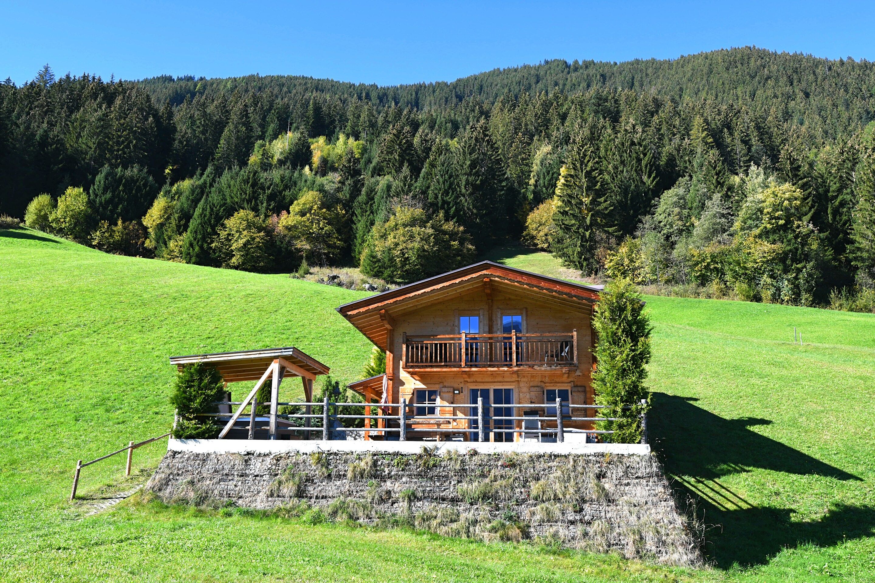A wooden chalet with a balcony, set on a green hillside with a forested background.