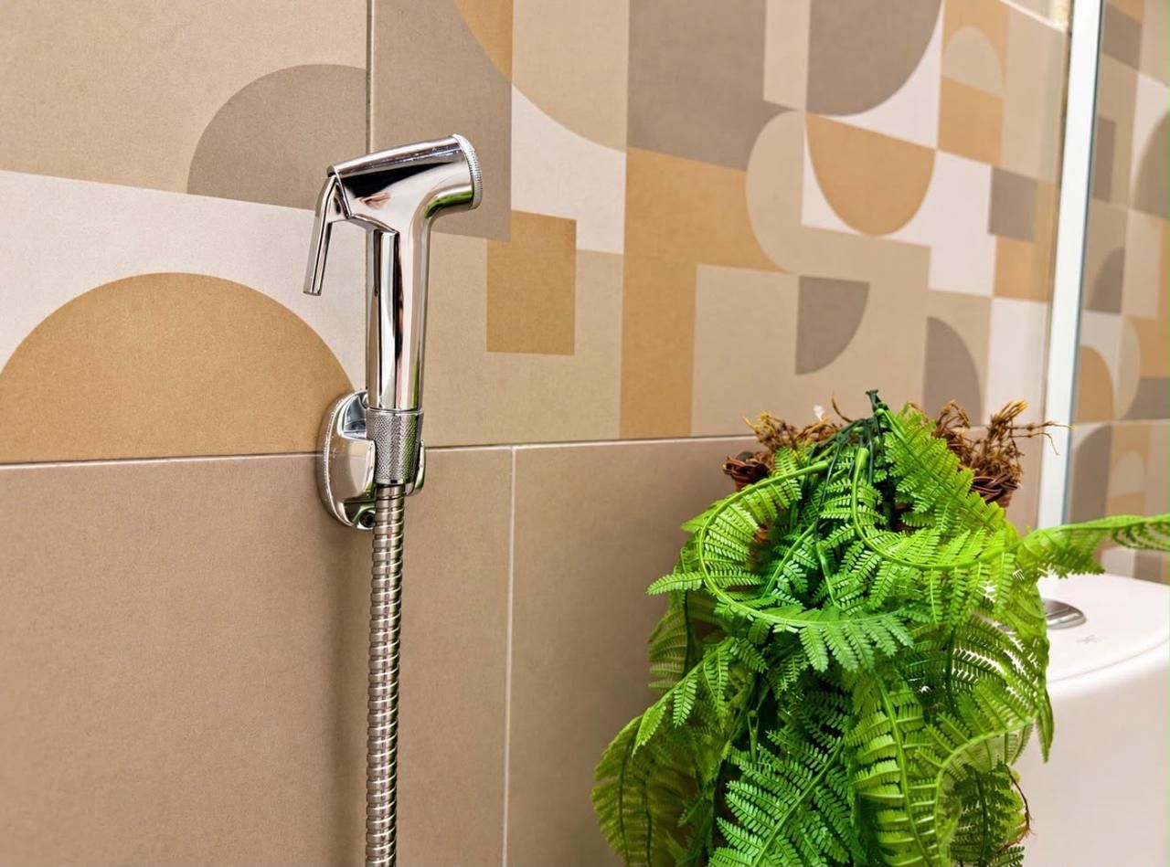 A bathroom with a shower head mounted on a tiled wall, a potted fern plant, and a toilet visible in the background.