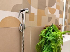 Une salle de bain avec une pomme de douche fixée à un mur carrelé, une fougère en pot et des toilettes visibles au fond.