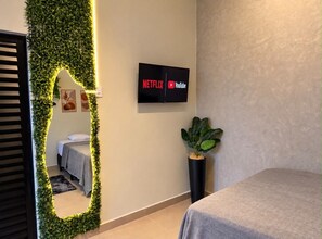 Une chambre d’hôtel avec un lit, un téléviseur fixé au mur affichant les logos de Netflix et YouTube, un miroir avec un cadre vert orné de motifs de feuilles, et une plante en pot.