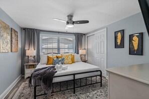 Een slaapkamer met een bed, nachtkastjes, een plafondventilator, een raam met jaloezieën en kunstwerken aan de muur.