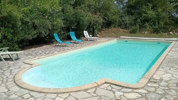 Una piscina rectangular con borde de piedra, rodeada por un patio de piedra. Hay dos sillas reclinables, una azul y una blanca, ubicadas cerca de la piscina.