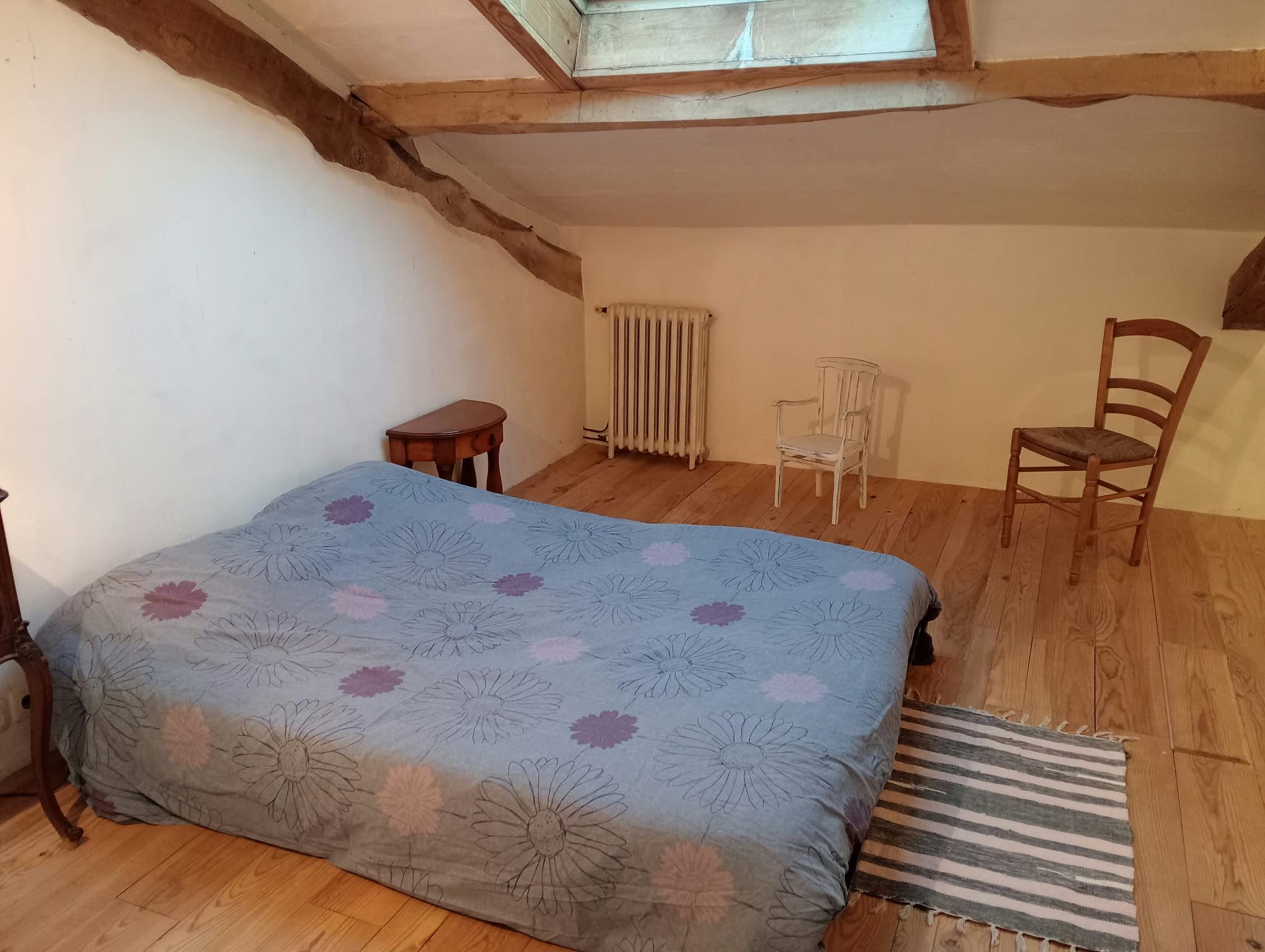 Une petite pièce chaleureuse, avec un plancher en bois, un lit recouvert d’une courtepointe à motifs floraux, un radiateur, une petite table, une chaise et une fenêtre avec un plafond orné de poutres en bois.
