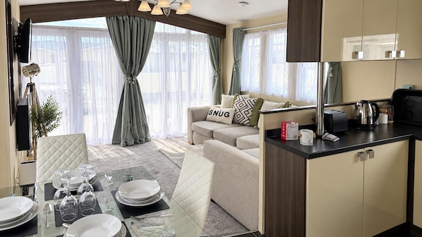 A modern living room with a sofa, a dining area with a glass table set, and a kitchen area with a coffee maker. - Abergele 2-bed Cabin, Sleeps 6 With Parking (Abergele)