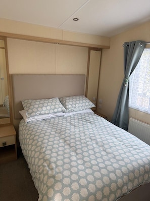 A single bed with a patterned bedspread, a wooden headboard, a nightstand, and a window with curtains.