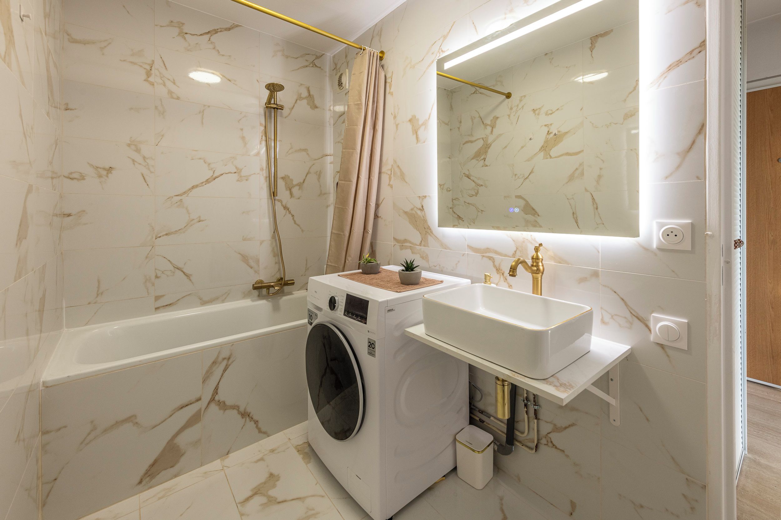 A modern bathroom with a marble wall, a bathtub, a washing machine, and a sink with gold fixtures.
