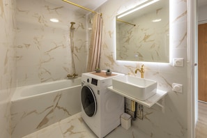 A modern bathroom with a marble wall, a bathtub, a washing machine, and a sink with gold fixtures. - Bright apartment near the beaches of Juan-les-Pins (Antibes)