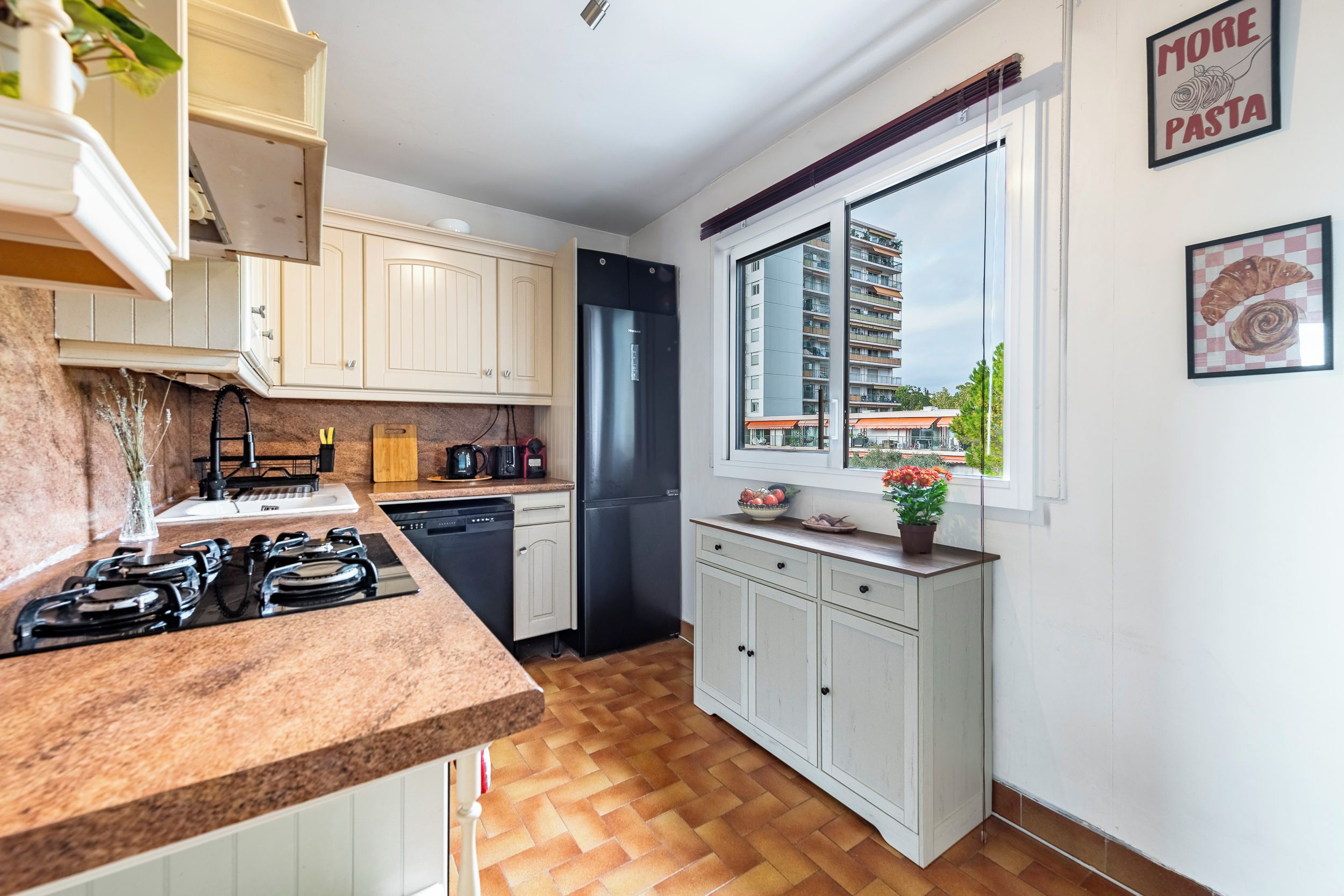 A modern kitchen with a built-in stove, a refrigerator, and a window with a city view.
