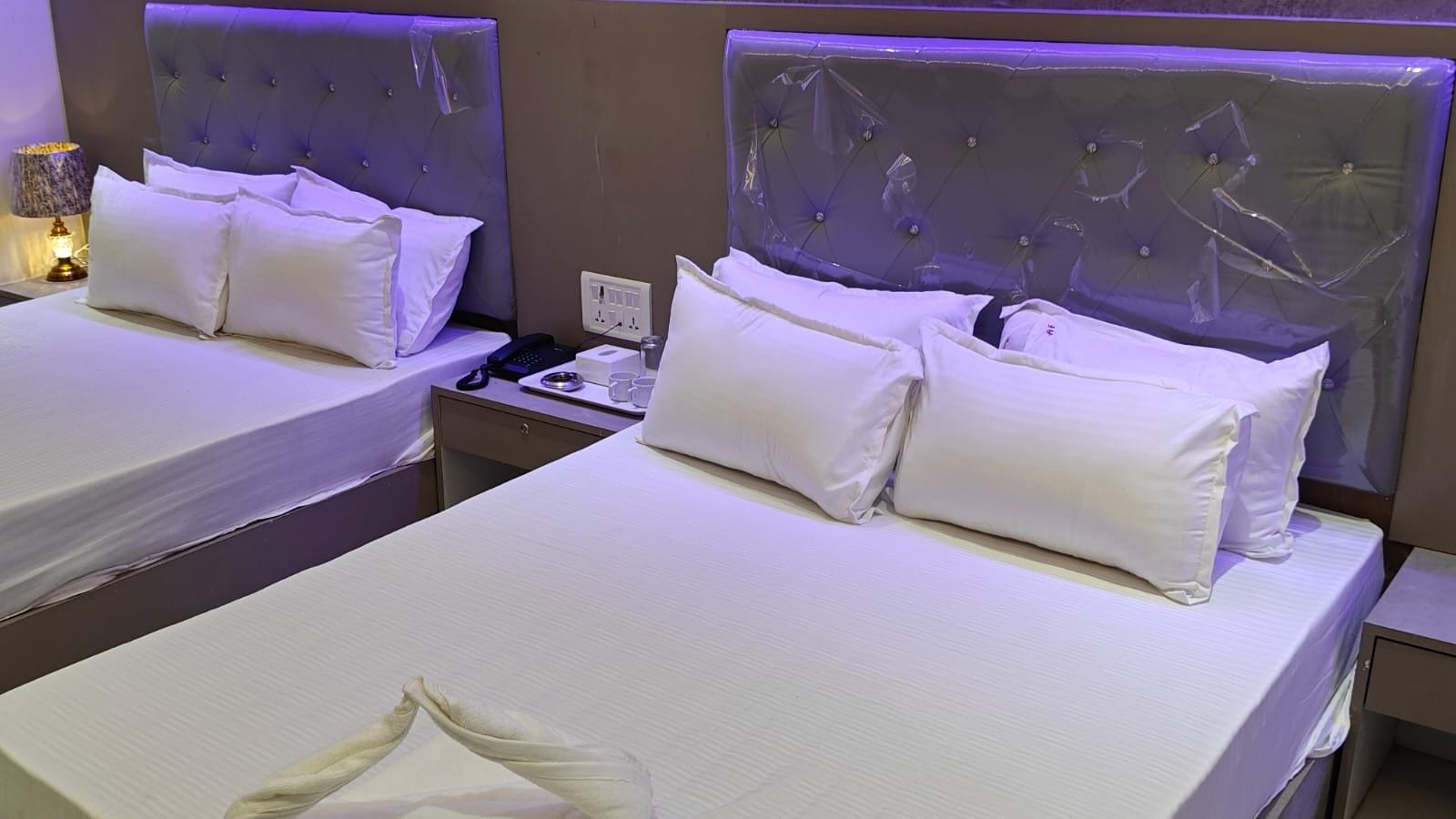 Habitación de hotel con dos camas, cada una con ropa blanca y almohadas, una mesita de noche con lámpara y un enchufe.
