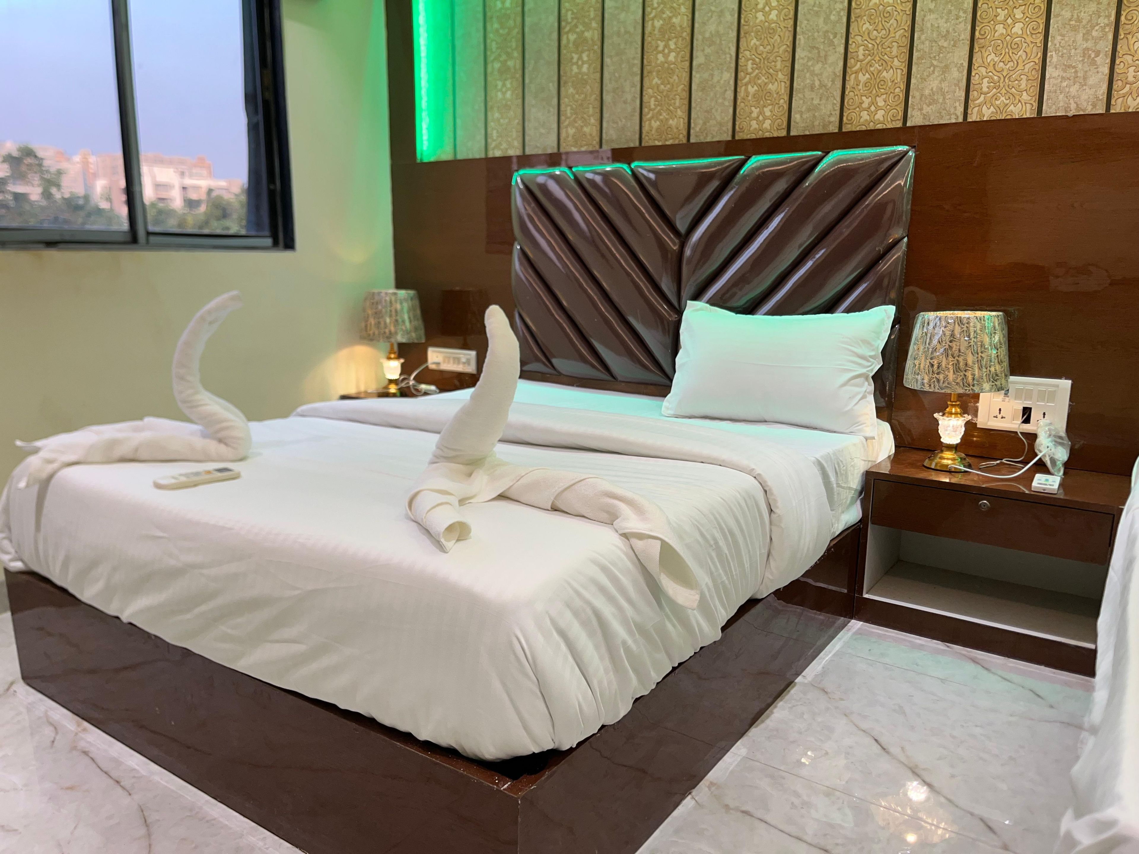 Habitación de hotel con una cama grande, cabecera con diseño geométrico, mesita de noche con lámpara y vista a edificios por la ventana.