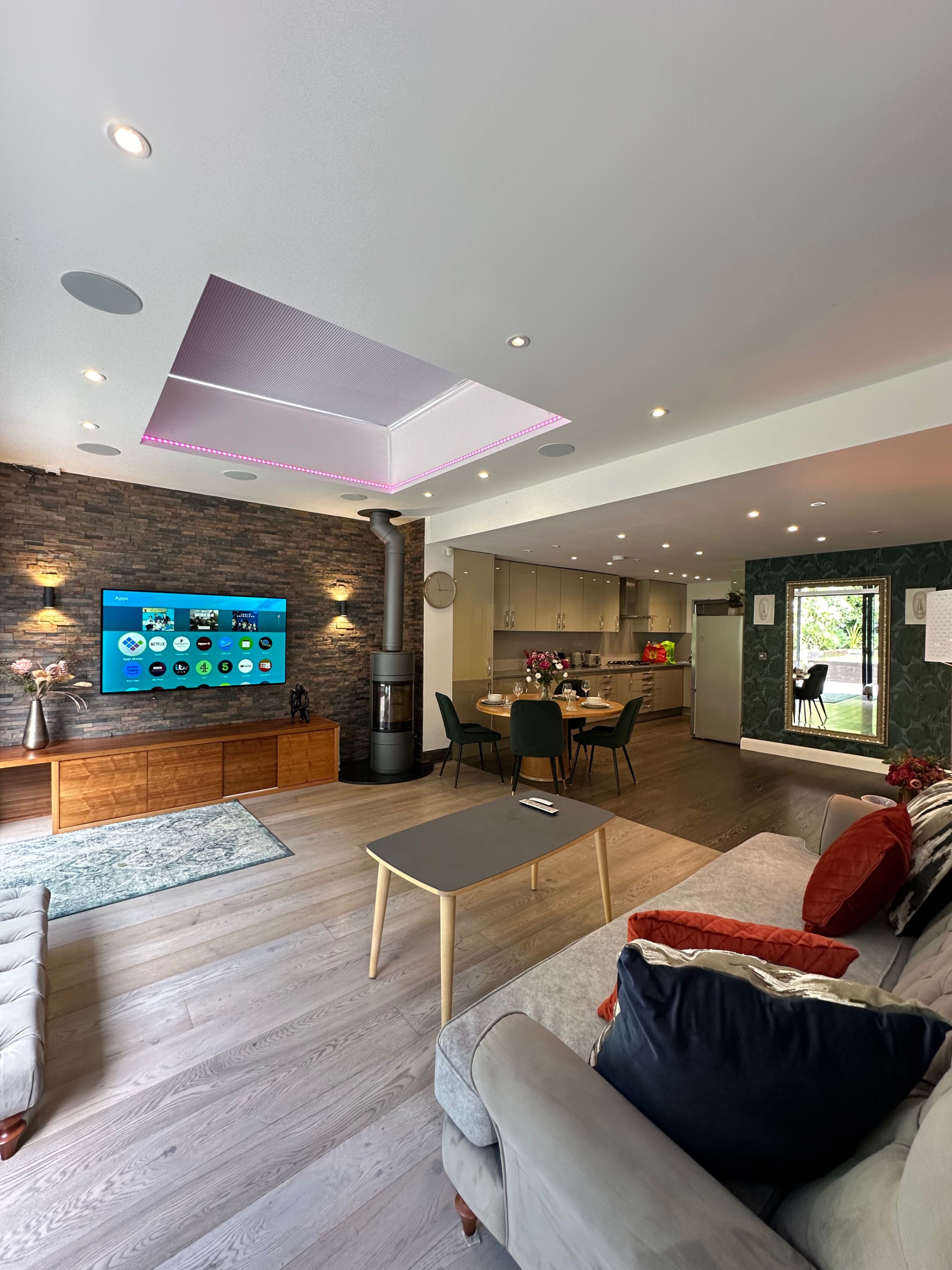 A modern living room with a flat-screen TV, a fireplace, a dining area, and a kitchen in the background.