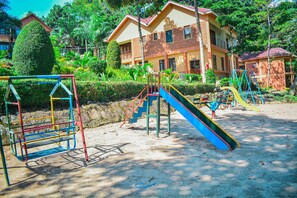A playground with slides and swings, surrounded by a building with a red roof and a well-maintained garden.