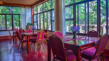 A dining area with a long table, multiple chairs, and large windows offering a view of greenery outside.
