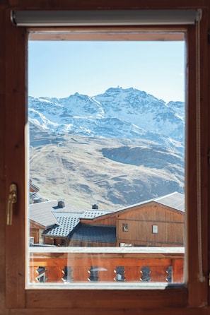 A wooden-framed window with a view of snow-covered mountains and a building with a sloped roof. - Balcons 647 (Les Belleville)