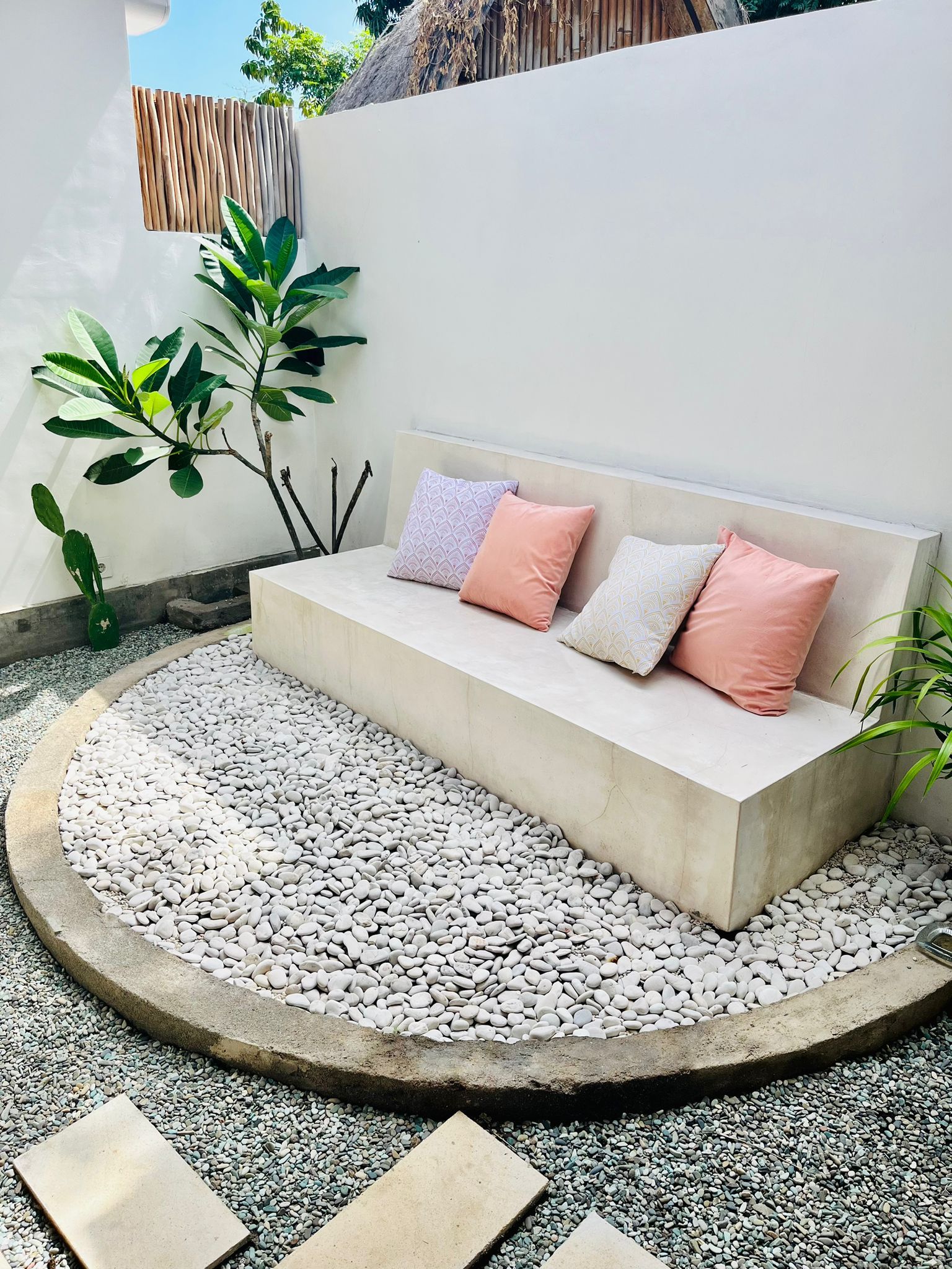 A concrete bench with cushions, surrounded by a gravel area, with a plant and a bamboo fence in the background.