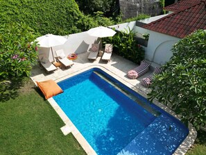 A rectangular swimming pool with a tiled edge, surrounded by a white wall and a red-tiled roof. There are lounge chairs and umbrellas, and a grassy area with a small orange cushion. - Villa Mila Gili Air (Pemenang Kabupaten Lombok Utara)
