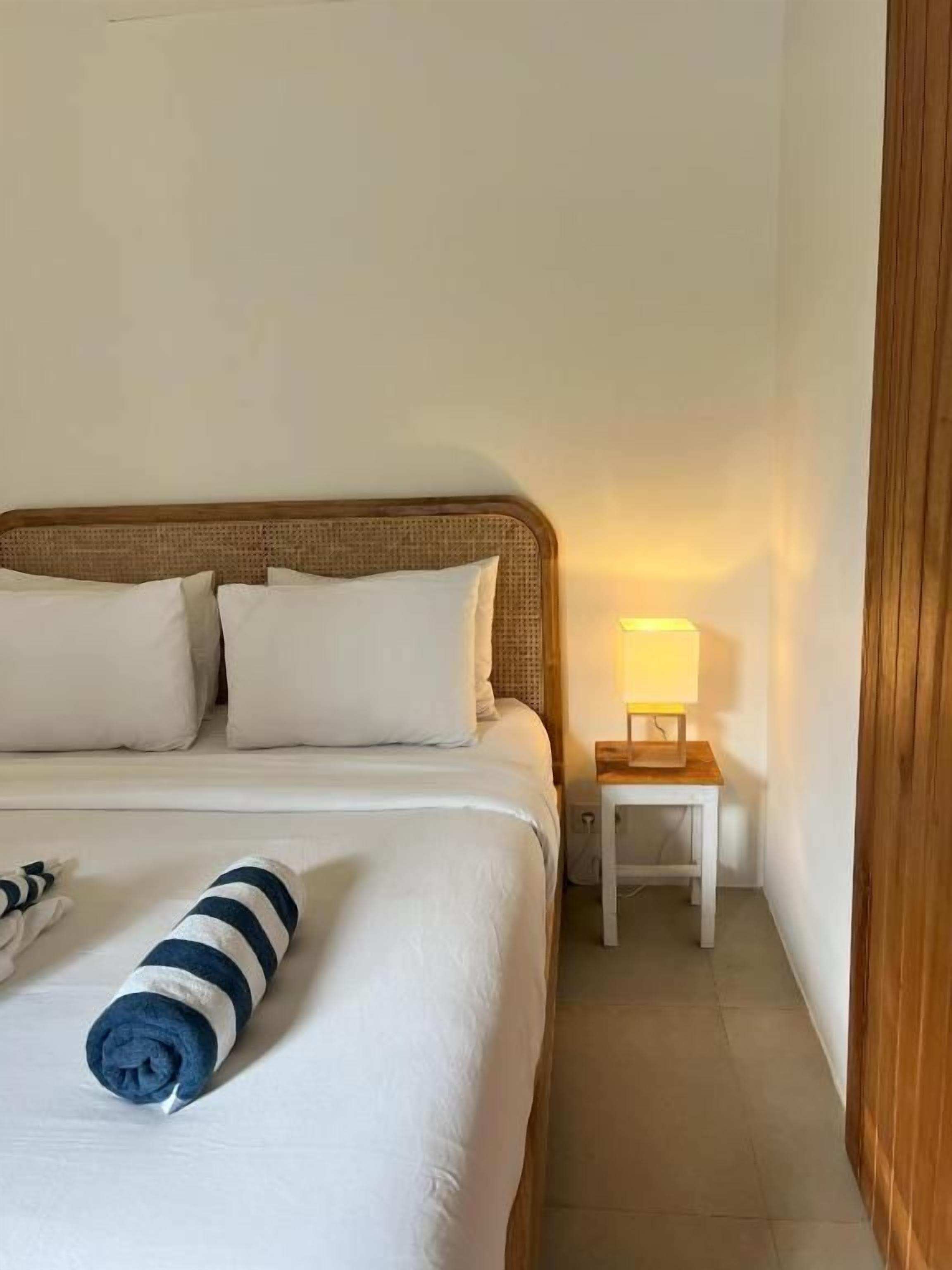 A neatly made bed with white bedding and a rolled towel, a bedside table with a lamp, and a wooden door.