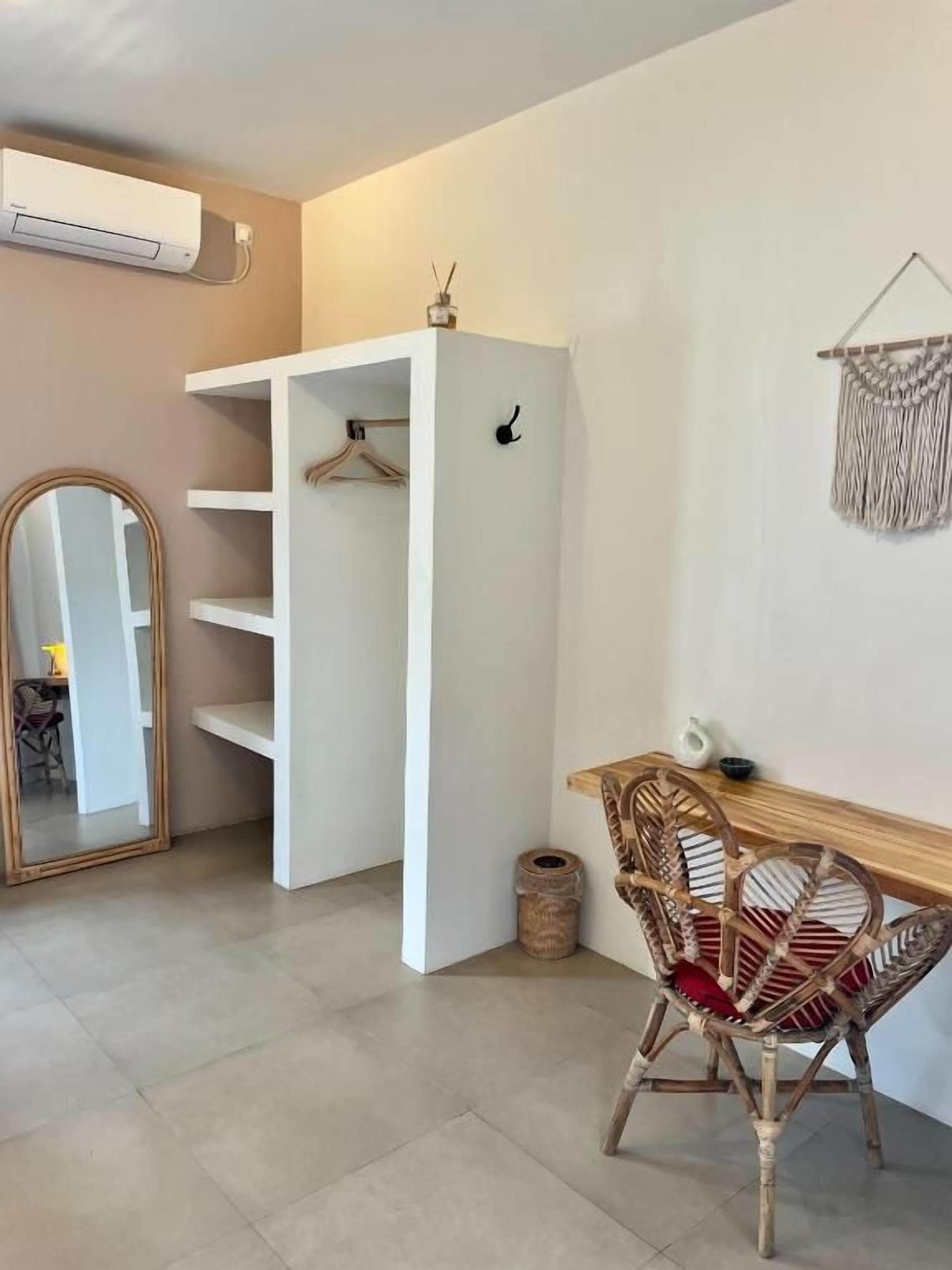 Una habitación con un armario blanco, una mesa de madera, sillas de mimbre, un espejo y un tapiz de macramé en la pared.