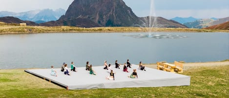 Un grupo de personas sentadas en un escenario frente a un lago, con montañas al fondo.