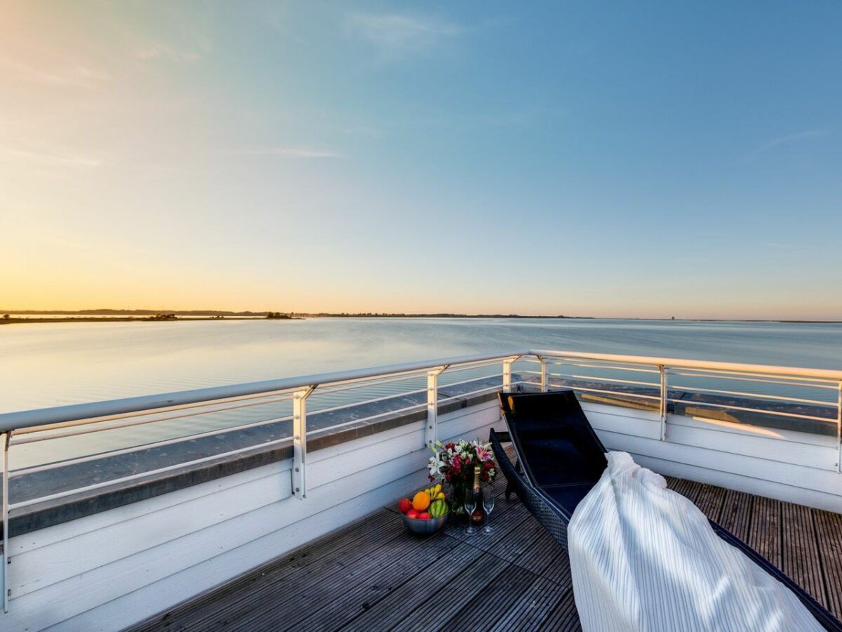 Una terraza con barandilla, una mesa con flores y fruta, y vista a una masa de agua tranquila.
