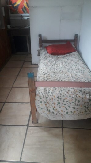 Quarto pequeno e estreito com uma cama, cabeceira de madeira e chão de azulejos.