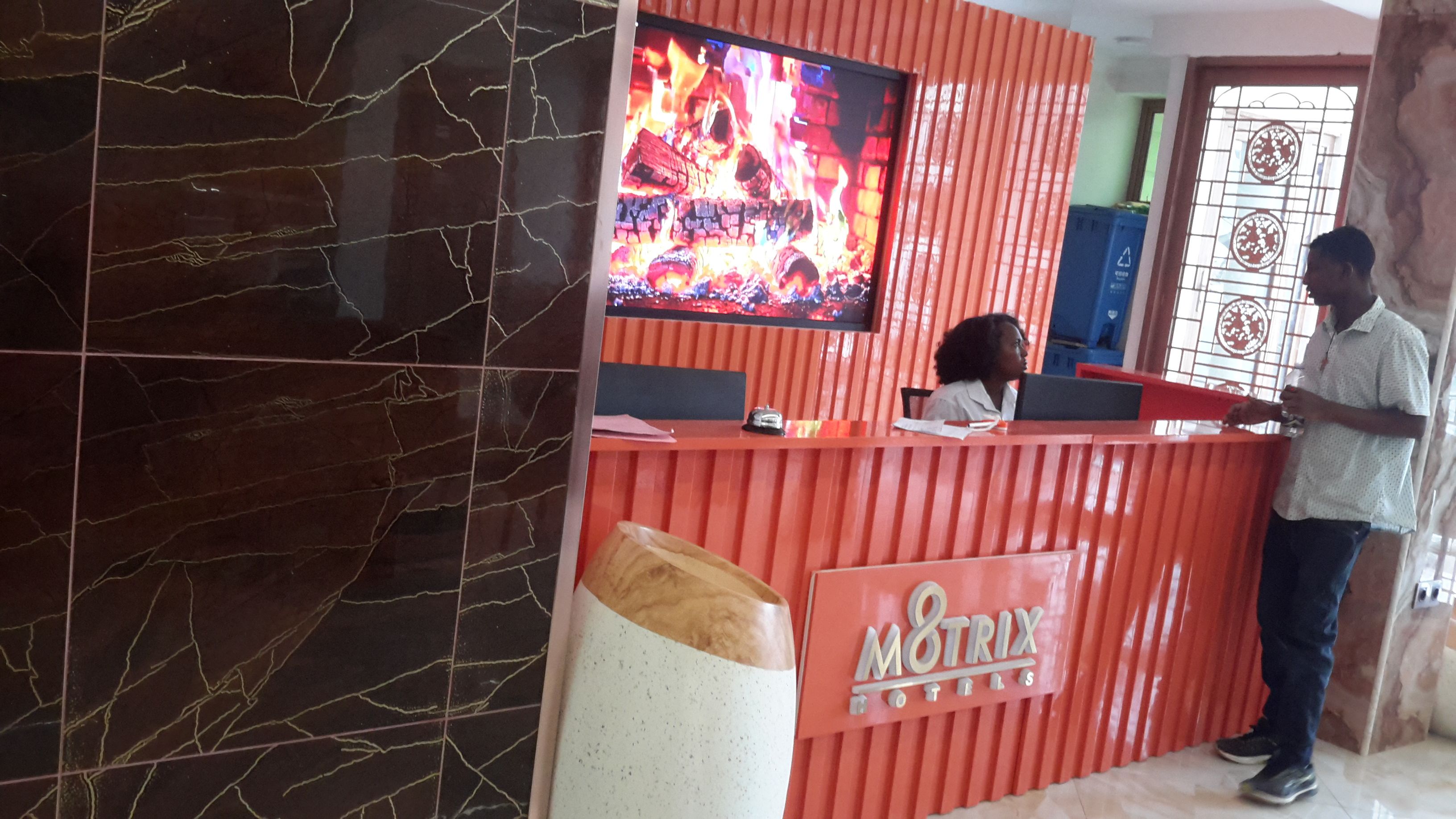 Un comptoir d’accueil avec un téléviseur diffusant une image de feu, une poubelle et une enseigne indiquant « MOTRIX HOTELS ».