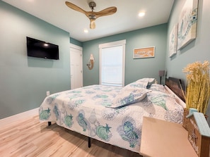 A bedroom with a bed, a ceiling fan, a TV, and artwork on the walls.