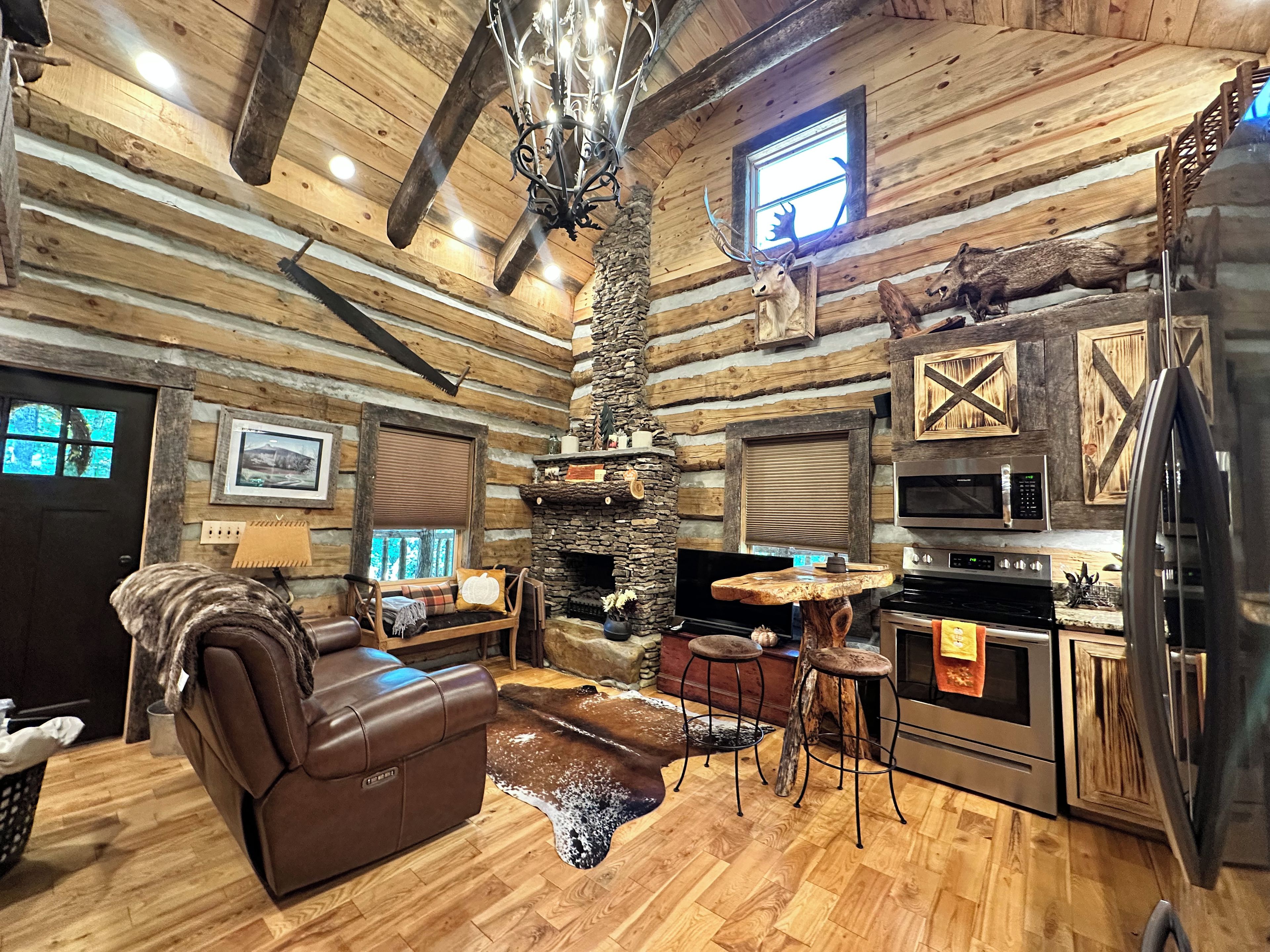 A log cabin interior with a fireplace, a leather sofa, a dining table, and a kitchen area.