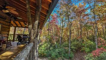 A rustic cabin with a wooden deck, a dining area, and a view of a forest with autumn foliage.