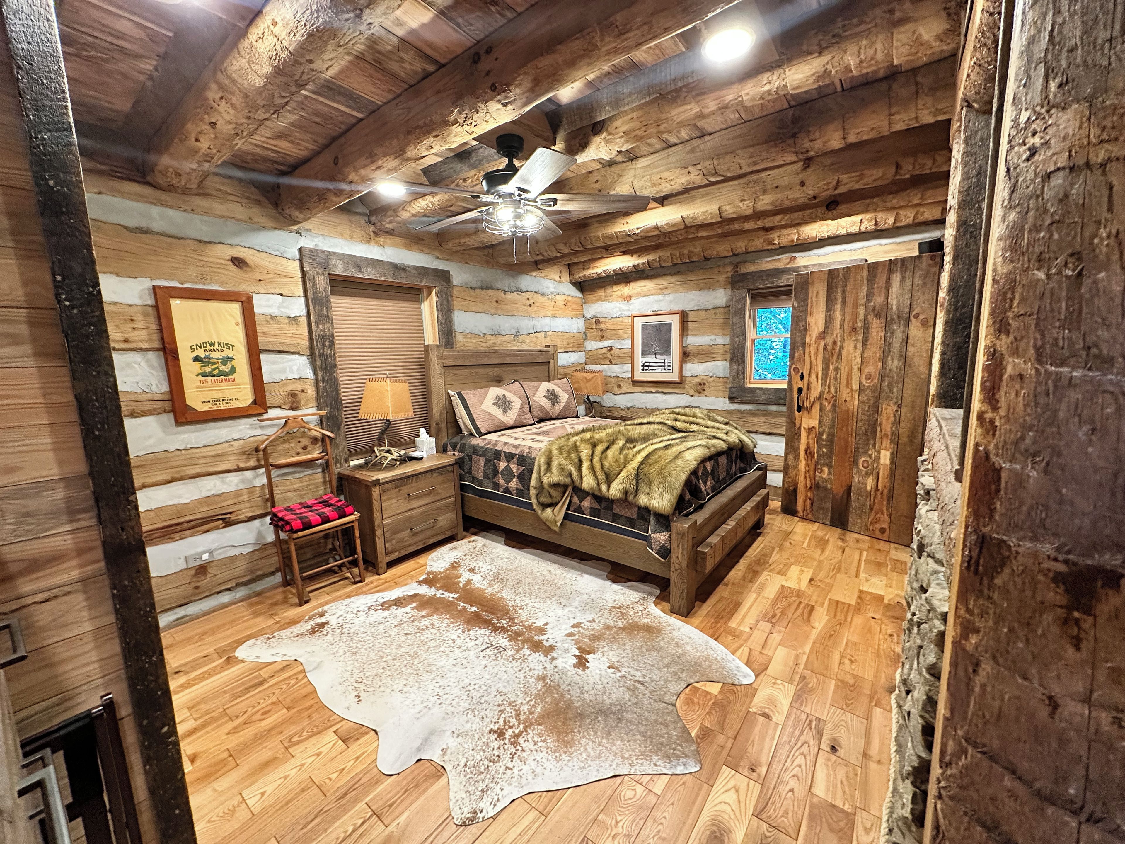 A log cabin-style bedroom with a bed, a wooden nightstand, a lamp, a framed picture, and a ceiling fan.