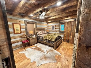A log cabin-style bedroom with a bed, a wooden nightstand, a lamp, a framed picture, and a ceiling fan. - Treehouse cabin Cozy 2-bedroom in Dobson, NC (Dobson)