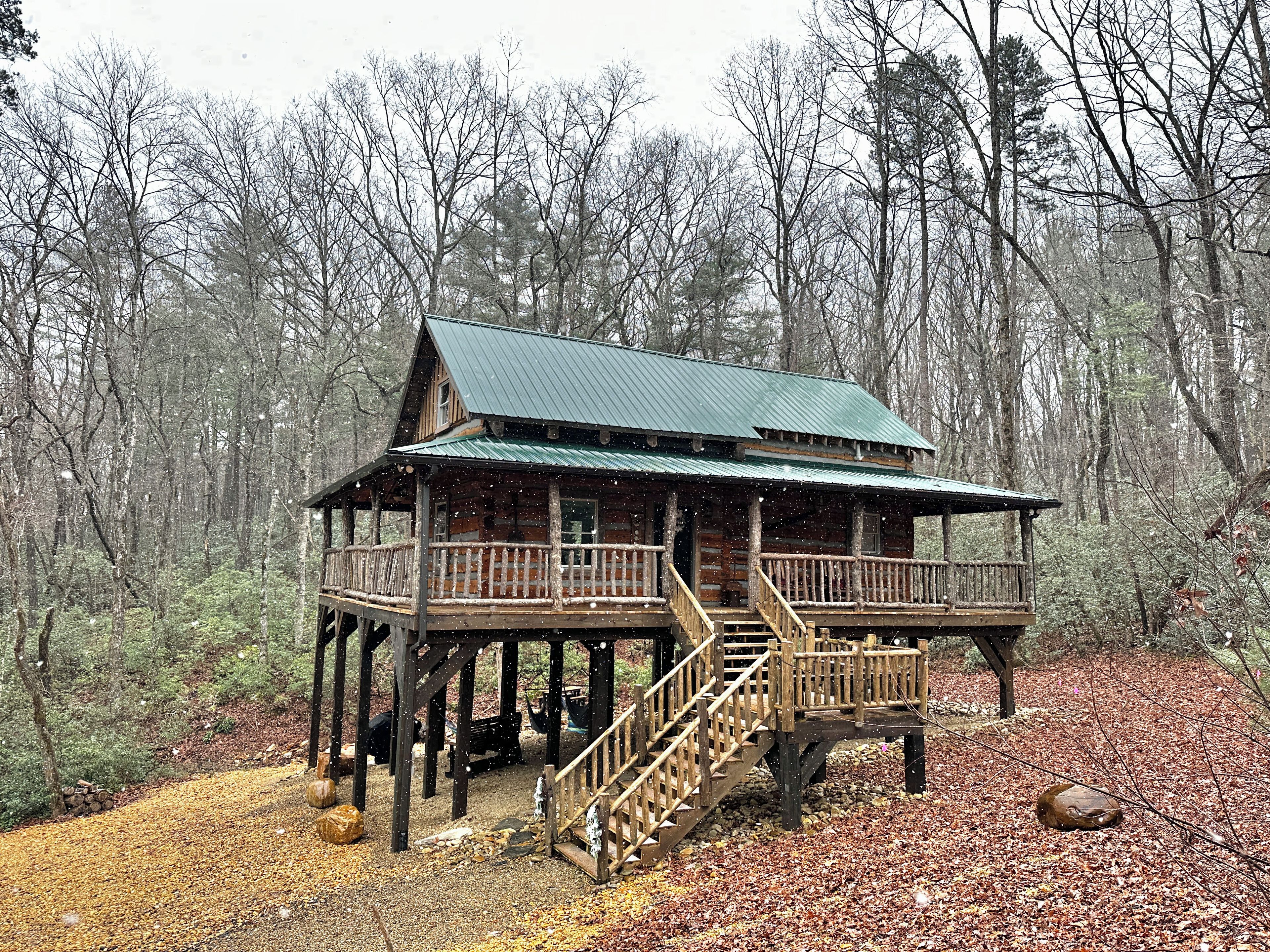 A log cabin with a balcony and stairs, surrounded by a forest.