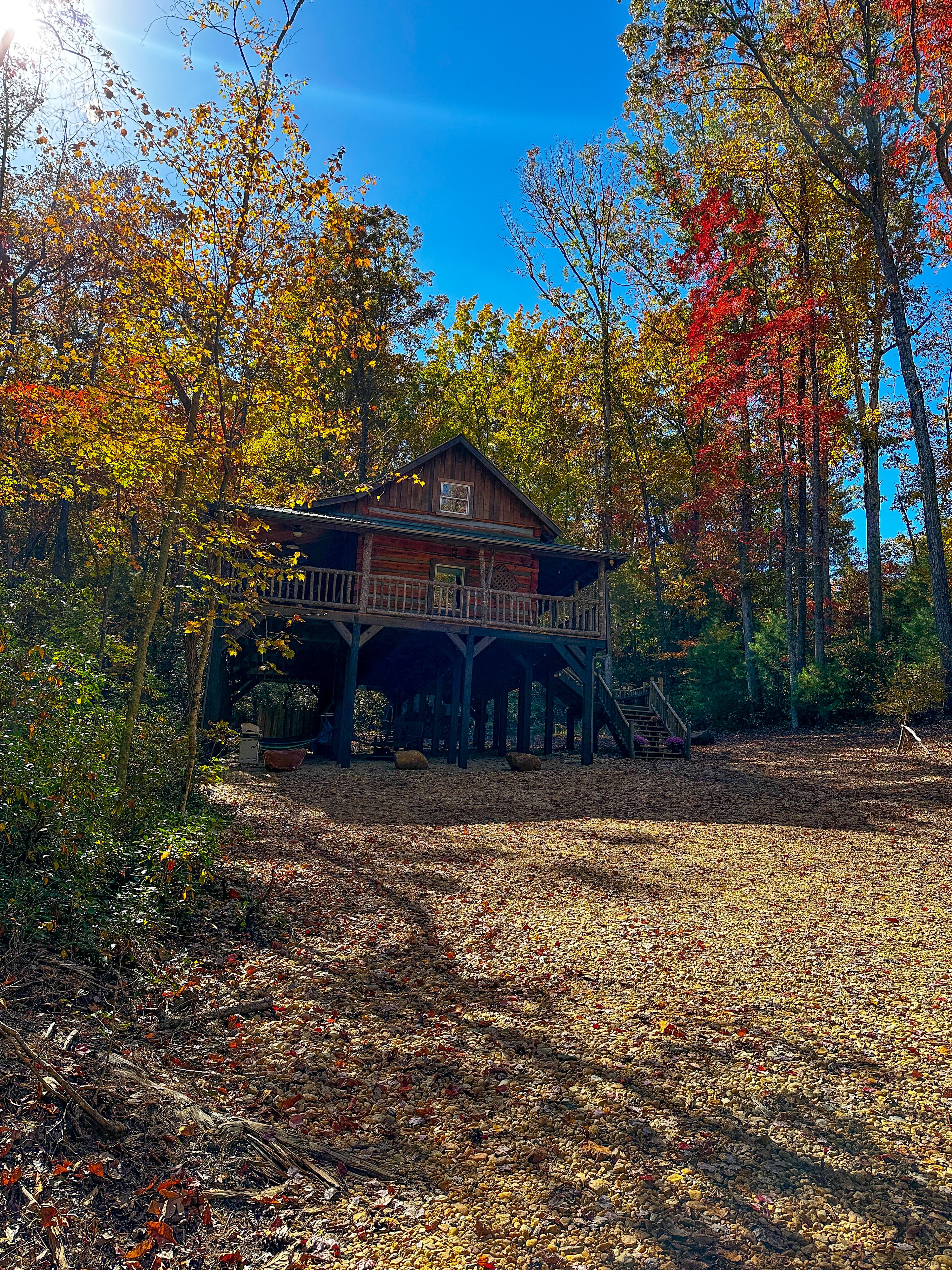 A rustic cabin with a wooden deck and a covered porch, surrounded by autumn foliage.
