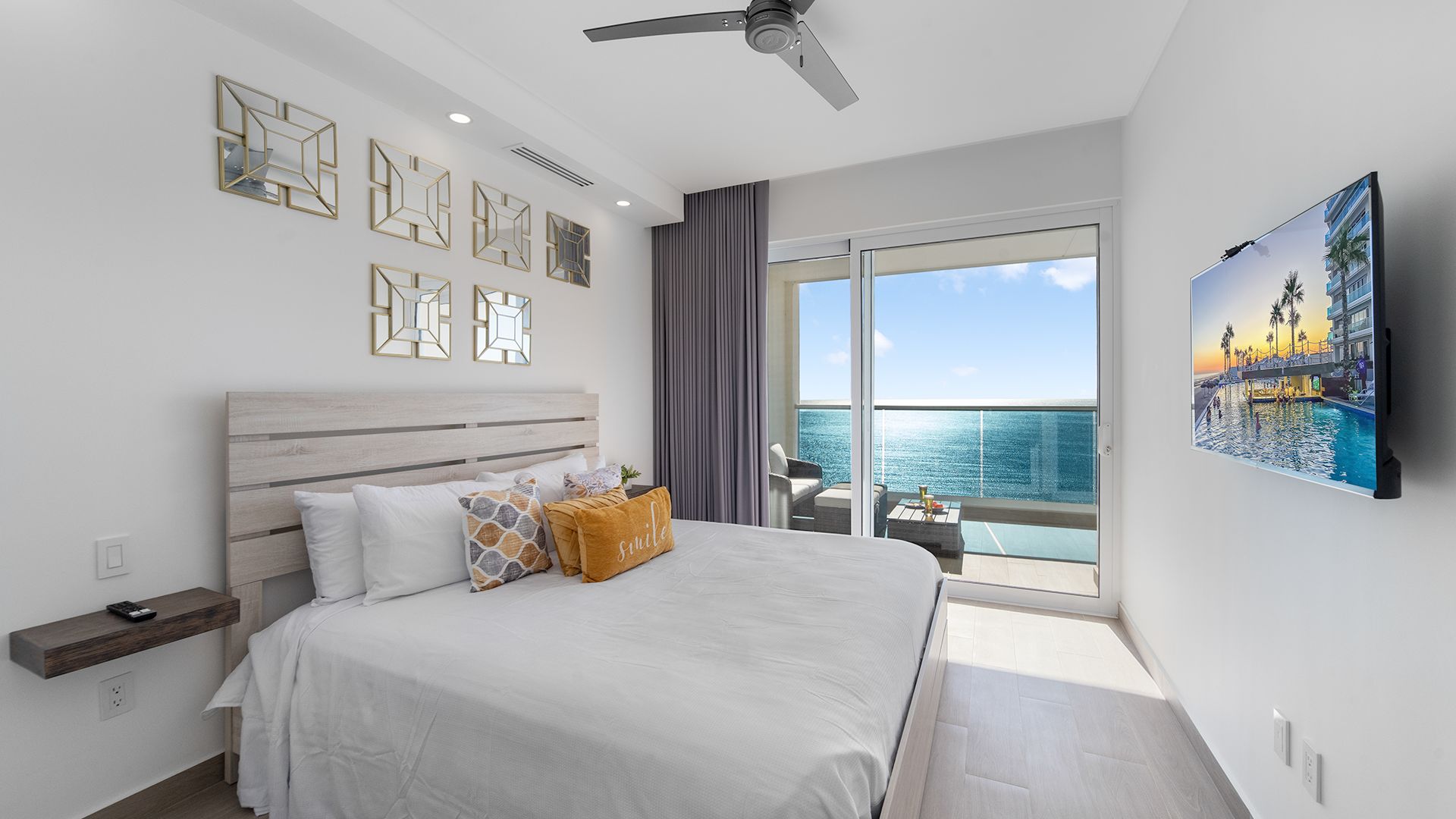 Een hotelkamer met een groot bed, een plafondventilator, een balkon met uitzicht op zee en kunst aan de muur.