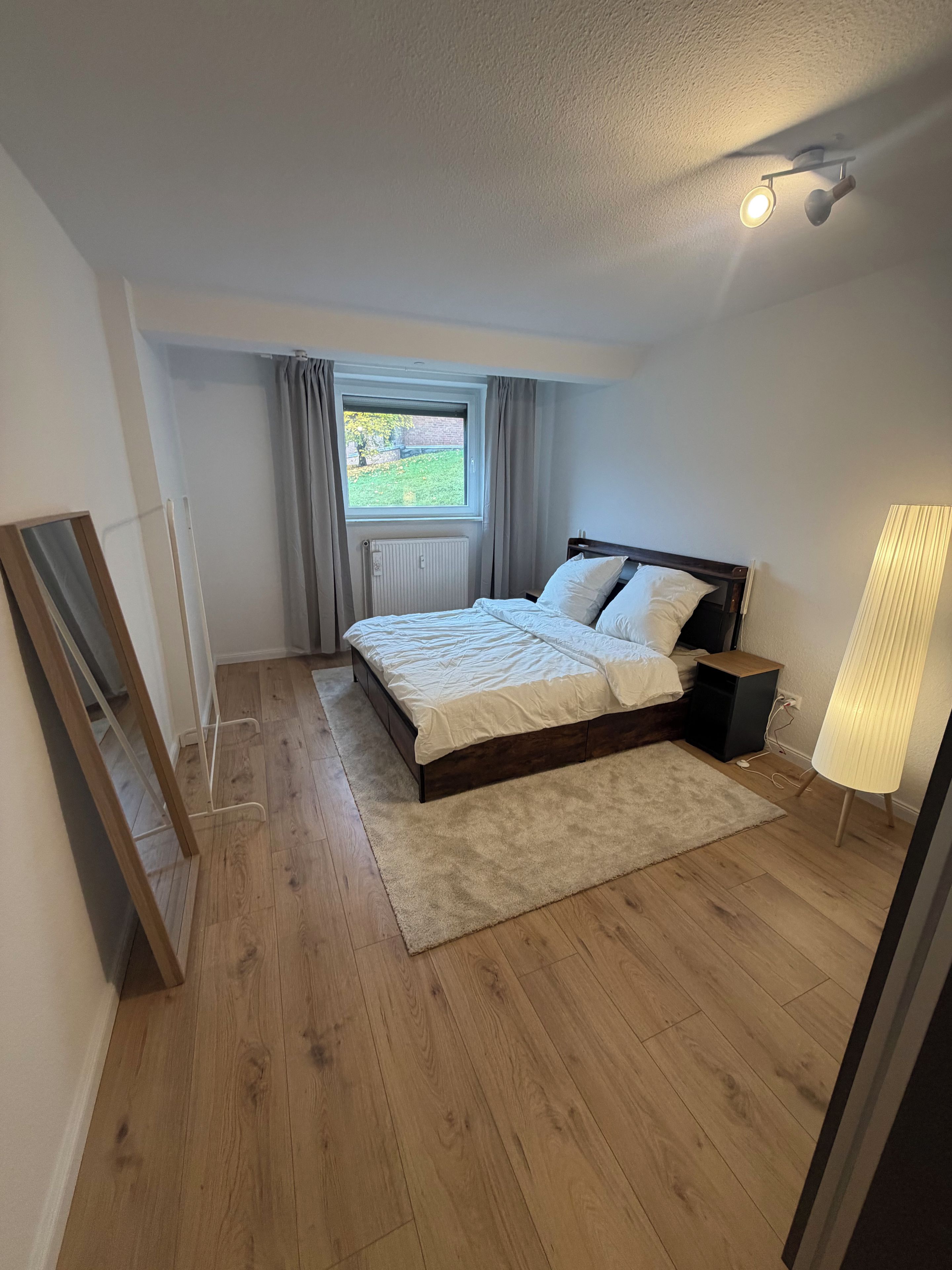 Un dormitorio con suelo de madera, una cama con ropa de cama blanca, una mesita de noche, una lámpara, una ventana con cortinas y un radiador.