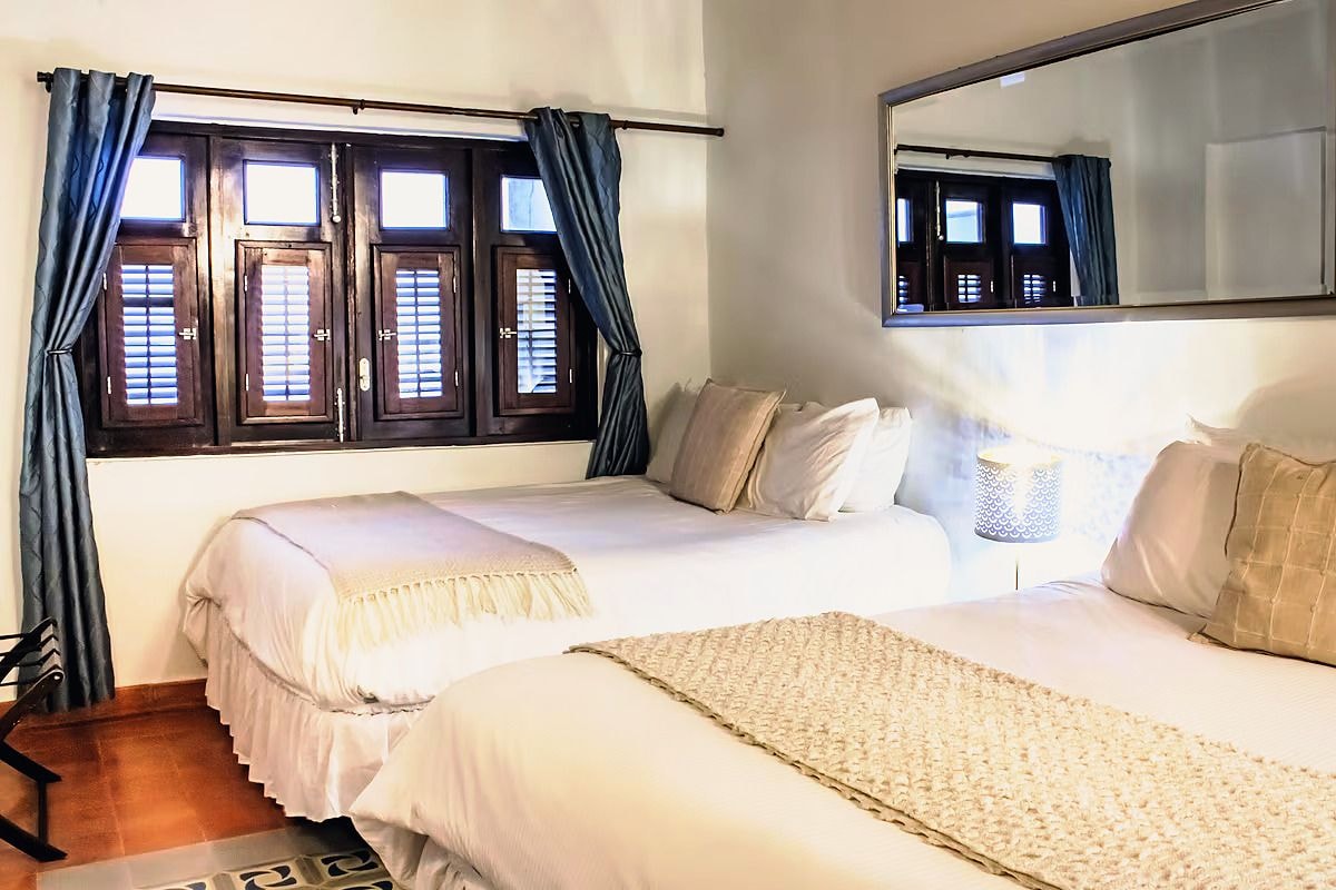 A bedroom with two beds, a window with shutters, a mirror, and a nightstand with a lamp.