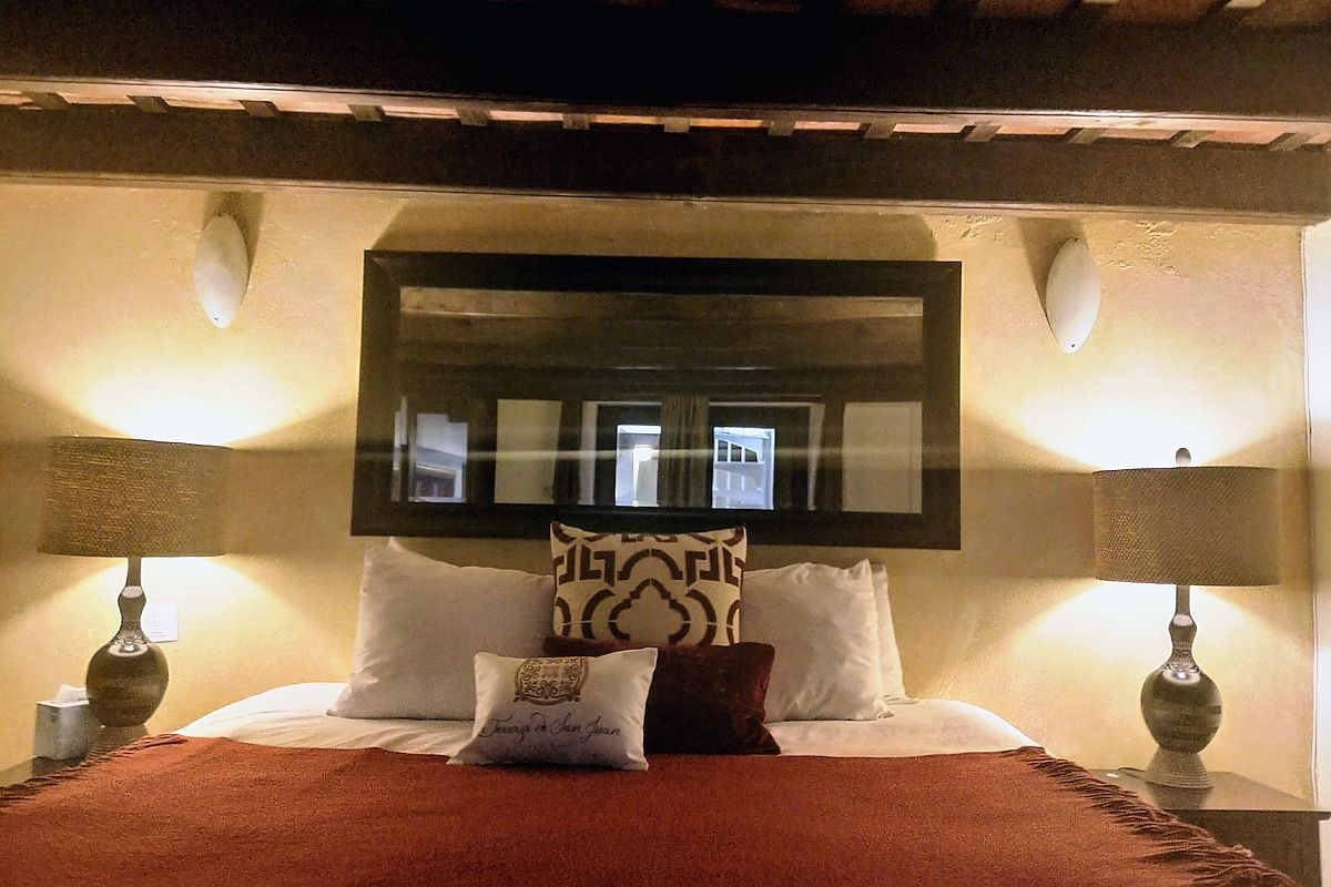A hotel room with a bed, two bedside lamps, a large mirror above the bed, and a patterned throw pillow.