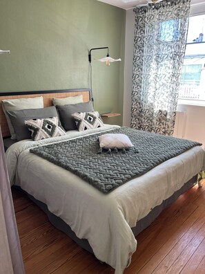 A neatly made bed with a patterned bedspread and pillows, a bedside lamp, and a window with patterned curtains. - New Listing (Royan)